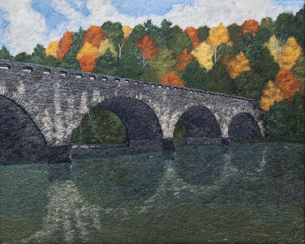 Felted wool artwork of an arched stone bridge over a river, with red, orange, yellow, and green trees in the background