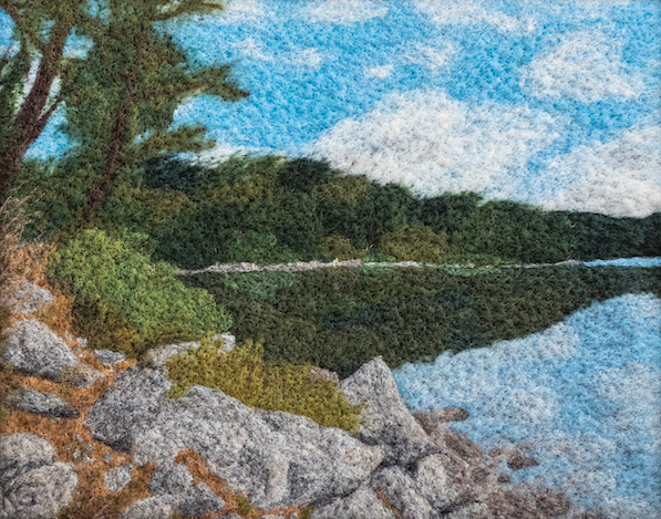 Felted wool artwork of rocks next to a lake that is reflecting the blue, partly-cloudy sky