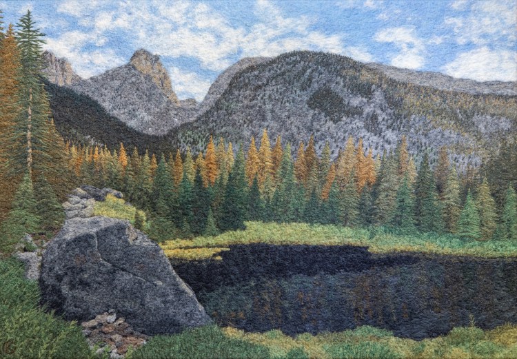 A felted wool artwork of a mountain landscape in the late afternoon. In the bottom third of the scene is a dark lake, surrounded by a meadow with a large boulder. On the far side of the lake is a stand of evergreen trees, lit by the sunlight. Above the trees are Rocky Mountain peaks.