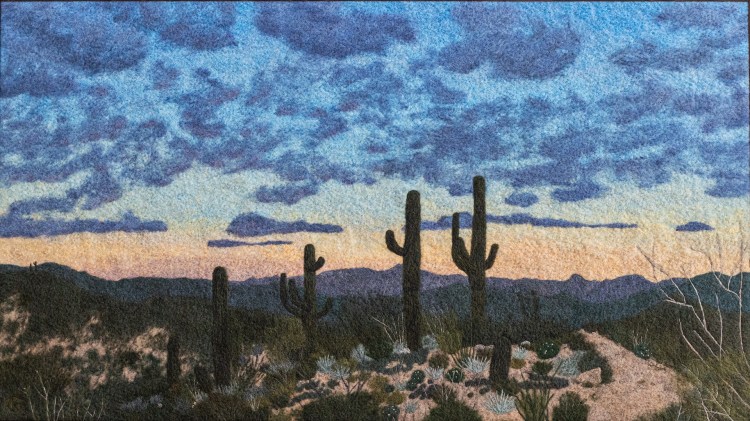 A felted wool artwork of featuring four saguaro cacti backlit against a colorful sky at sunset. Periwinkle clouds are overhead, desert vegetation is in the foreground, and a trail winds into the felting on the right side.