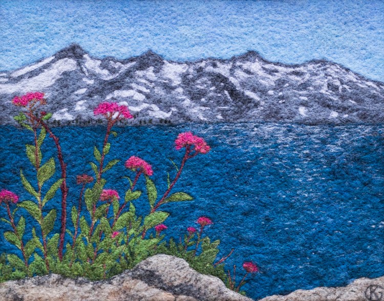 Felted wool artwork of pink flowers growing on a rock next to a blue mountain lake. At the far side of the lake is a gray ridge of snow-covered mountains.