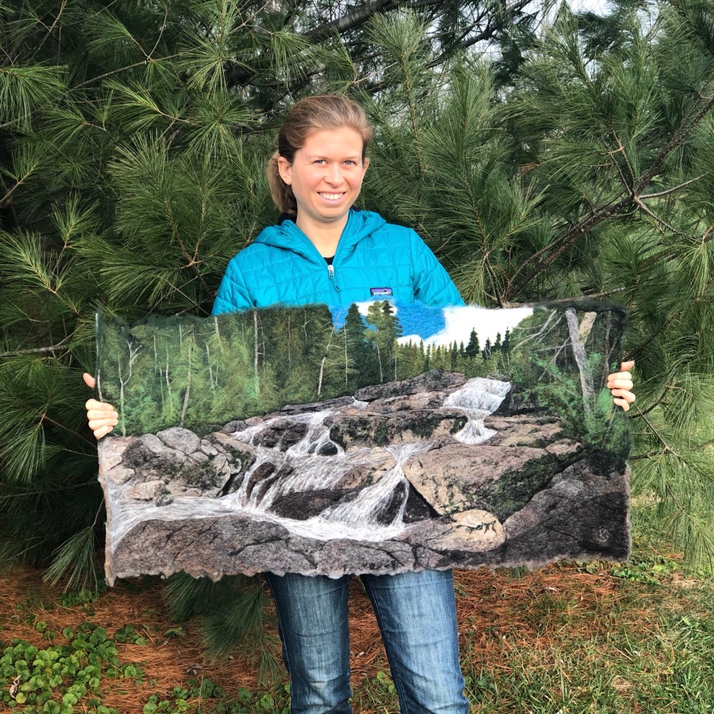 Artist Kris Grenier smiles while holding a wool felting of a waterfall in the Adirondacks