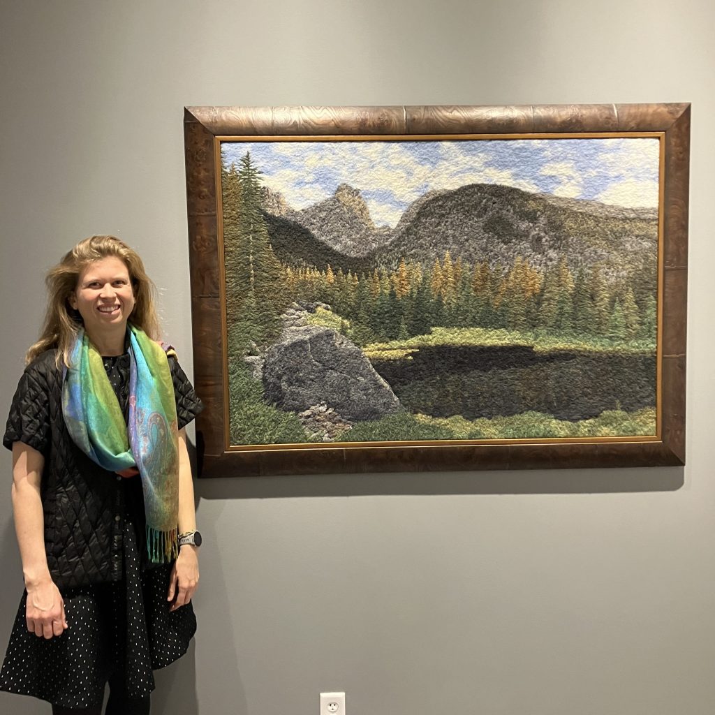 Artist Kris Grenier poses next to a framed woolen landscape of a mountain scene in Colorado
